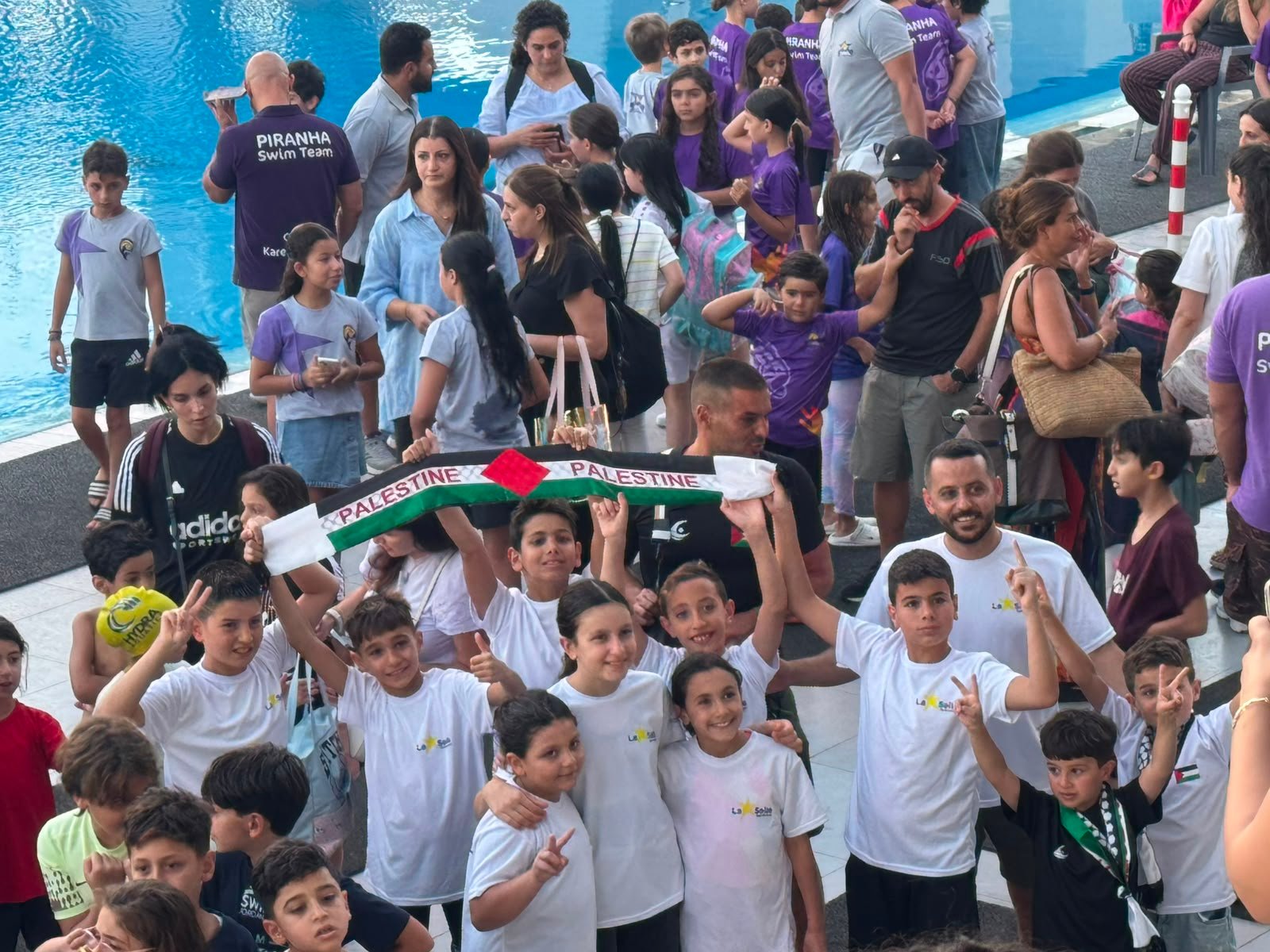 De La Salle swimmers, coaches, and families celebrating together beside the pool with a Palestine scarf.